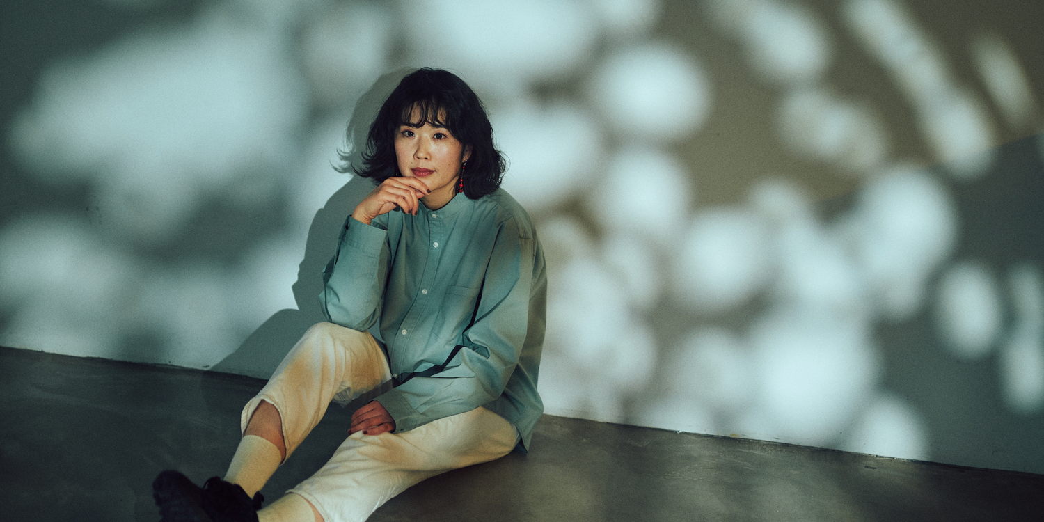 SOLITの服を着るモデルがコンクリートの壁の前で座り、カメラを見つめている