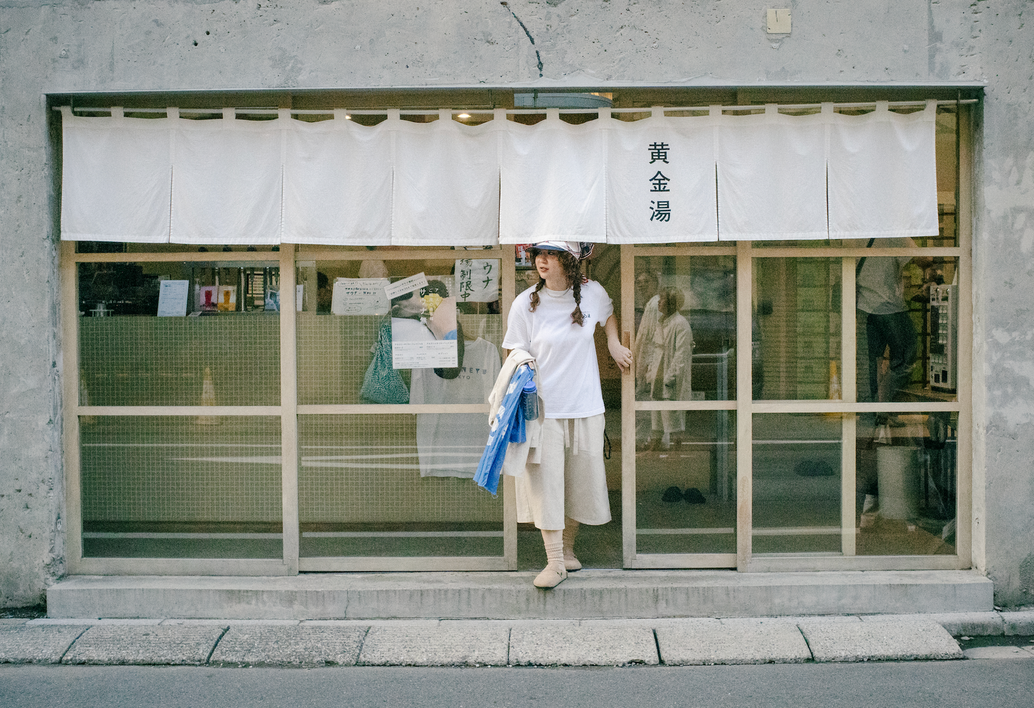 黄金湯という銭湯の入り口からでてくるパーマヘアの女性。麻のようなかろやかなコットンのセットアップを着用している