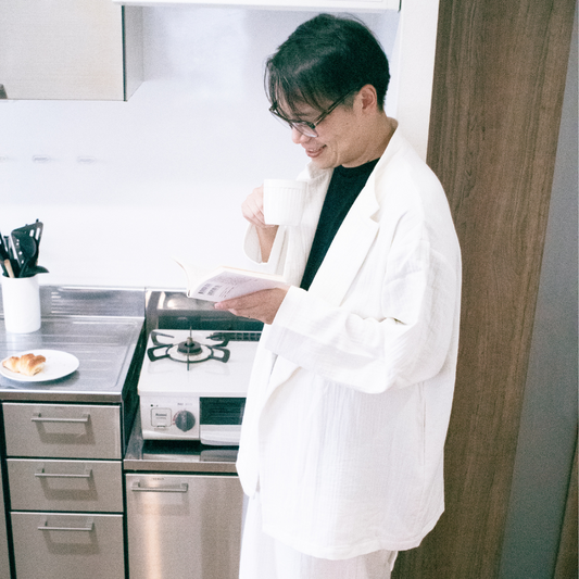 厚手の白いコットンのセットアップを着ている人がキッチンによりかかり、コーヒーを飲みながら片手で文庫本を読んでいる様子